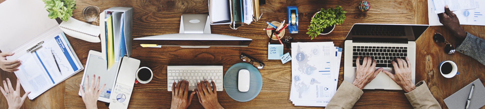 Image of various hands shown from overhead, working at notebooks, laptops, and with charts at a wooden table.