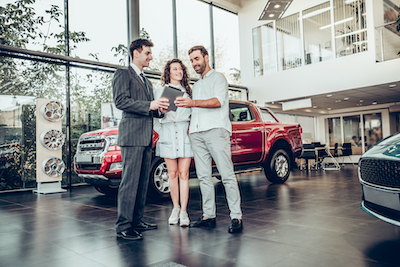 A man and woman standing in a dealership showroom, with a sales agent holding a tablet and closing a deal. A red truck is visible behind them.